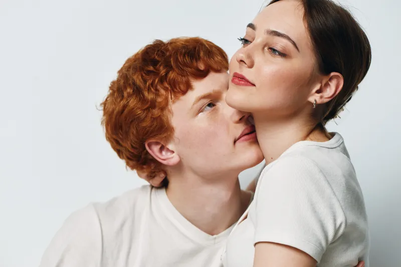 Couple, young man and woman embracing while sitting close together with affectionate expression on white background in studio. Intimate moment of love and connection between two people.