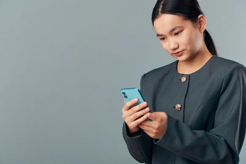 Woman holding smartphone with both hands looking at screen focusing on communication or message. Casual business outfit with dark hair pulled back and neutral background for technology concept.