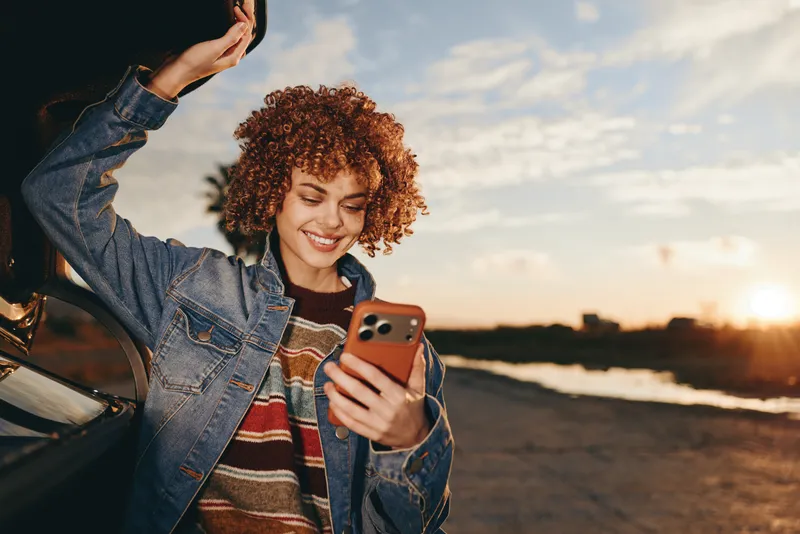 woman lifestyle, smiling with curly hair, denim jacket, holding smartphone for selfie by ocean sunset, outdoors on beach, warm light, relaxed pose, evening breeze