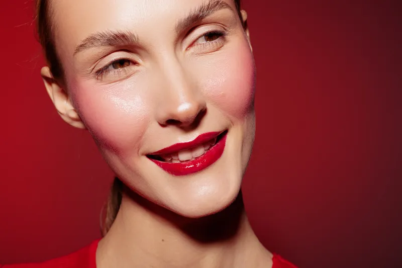 Close-up of smiling woman with red lipstick and glowing skin on red background. Concept of beauty, makeup, skincare, and confident female portrait with natural light and vibrant colors.