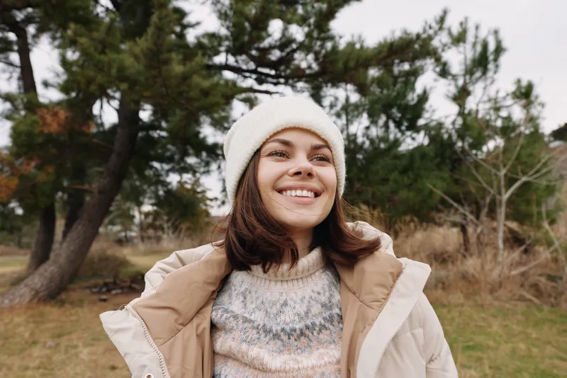 Woman smiling outdoors in a cozy knit sweater and beige jacket, standing in a park with trees, showcasing casual fashion, comfort and relaxed confidence.