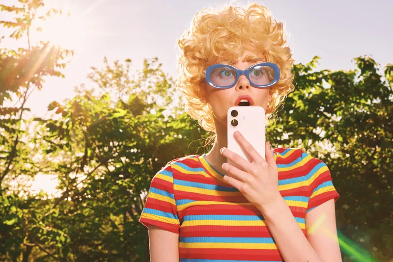 A stylish woman poses outdoors in a rainbow striped top and retro sunglasses, holding a smartphone. Bright sunlit scene conveys playful lifestyle, fashion, and carefree summer energy.