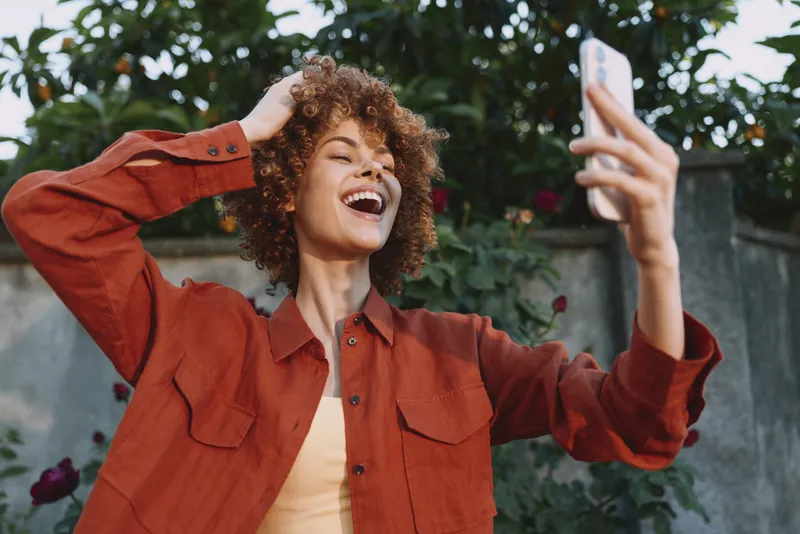 Selfie of a joyful young woman with curly hair wearing a rust orange jacket and light top, taken outdoors in a lush garden with blooming flowers