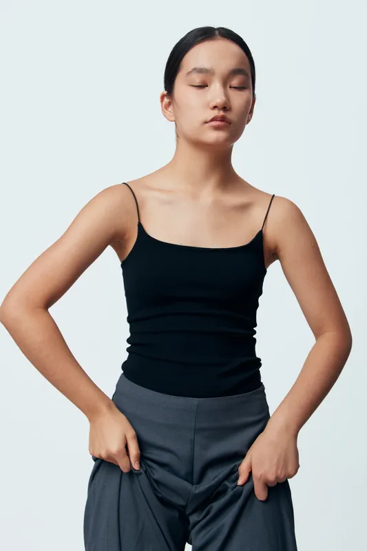 Young woman wearing black camisole and gray pants posing with eyes closed on white background. Minimalist fashion portrait with natural makeup and simple hairstyle in studio.
