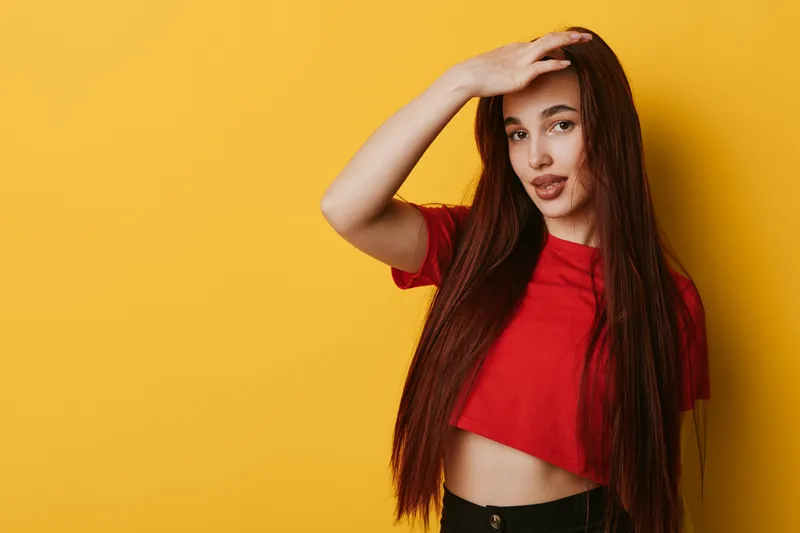 Woman with long hair wearing red cropped t shirt posing with hand on forehead on yellow background. Portrait of female model with casual style and confident expression.