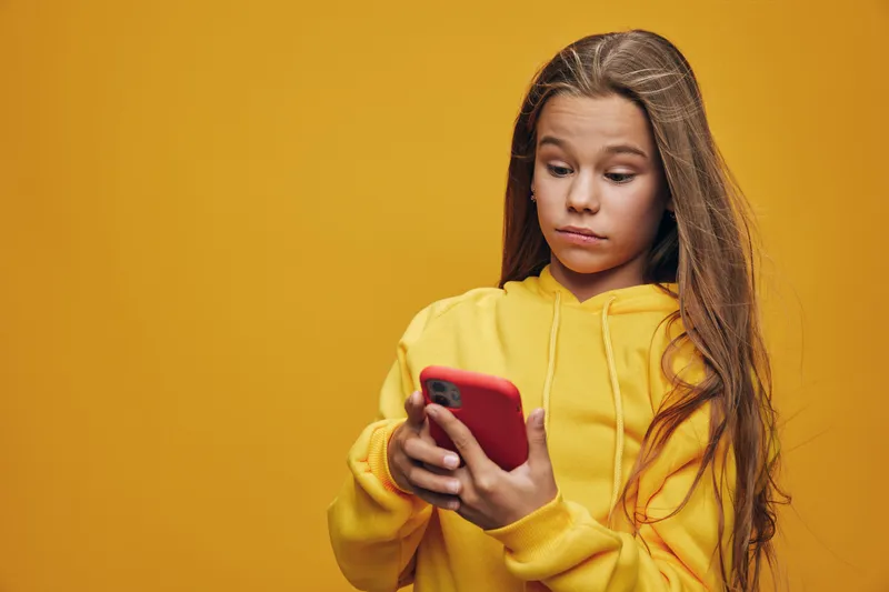 Girl in yellow hoodie looking at smartphone with focus on yellow background with casual style and long hair indoors for technology use and youth.
