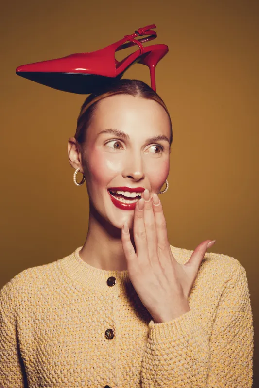 Young woman with red high heel shoe on her head smiles and covers mouth with hand. Studio portrait on brown background. Concept of fashion, fun, and playful mood.