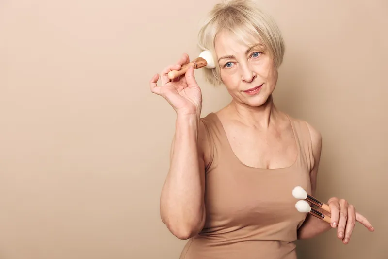 Mature woman with short blonde hair applies makeup using brushes on beige background. Concept of beauty, skincare, self-care, and natural aging with soft lighting and calm expression.