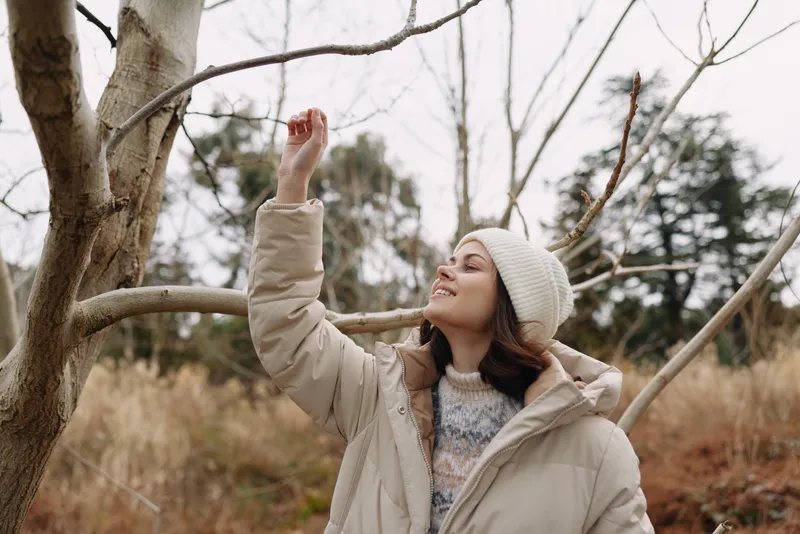 Woman in a beige coat and knit beanie reaches toward a bare branch in a park, capturing a calm winter moment amid autumn tones and soft light.