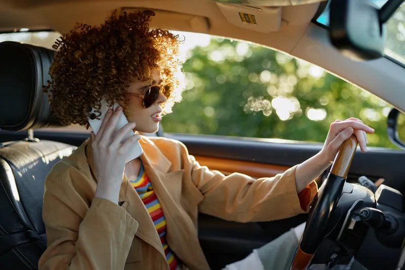 Woman driving a car, talking on a phone, wearing sunglasses and a blazer. Warm retro film color palette conveys casual urban lifestyle and spontaneous travel moments.
