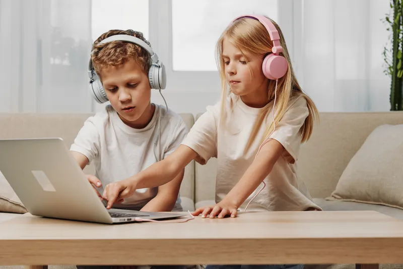 Two children with headphones use laptop together at home, focused on screen, sharing technology and digital learning concept.