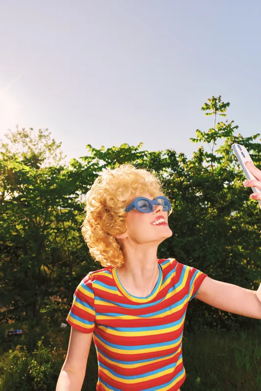 Smiling woman with curly hair taking a selfie outdoors in a sunny park, wearing a colorful striped shirt and sunglasses. Retro film color mood highlights lifestyle, happiness, and vibrant self