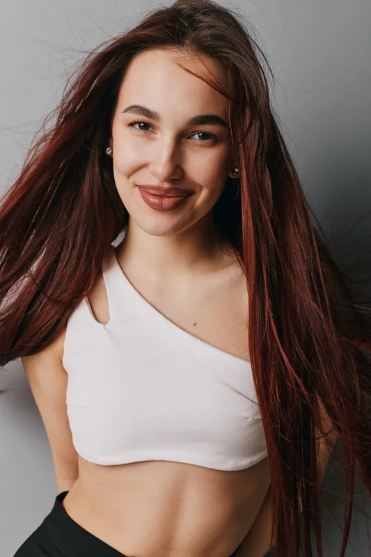 Young woman with long hair smiling and posing in white crop top with cutout detail against gray background, confident and happy expression in studio portrait.