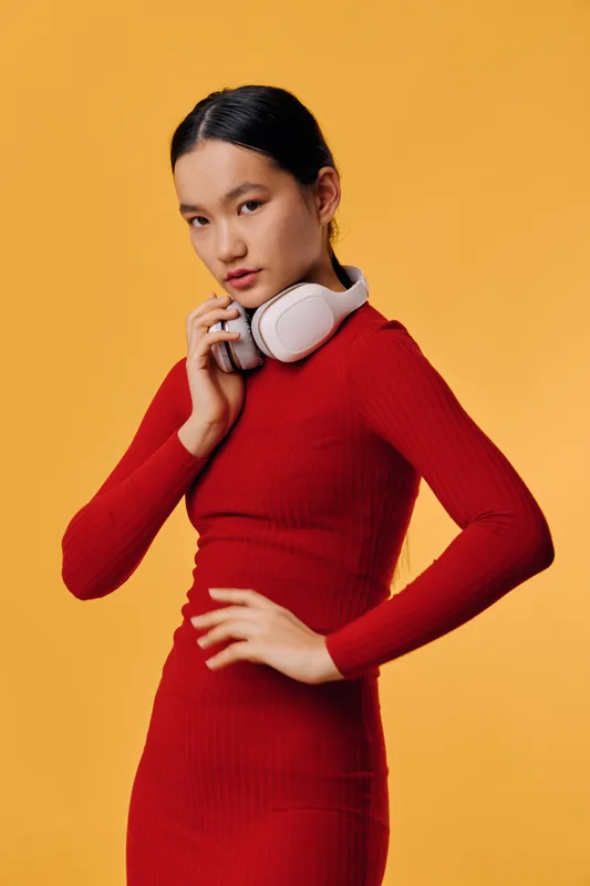 Young woman in red dress posing with wireless headphones around neck against yellow background, looking confident and stylish with hand on hip in studio portrait. Modern fashion and technology.