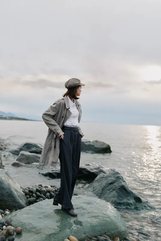 Stylish woman in long coat and cap stands on rocky shore looking at sea during cloudy day. Concept of fashion, autumn, outdoor lifestyle, and contemplation by water.