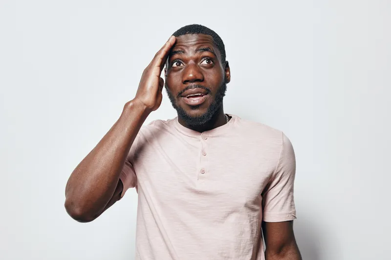 African American man with dark skin wearing light pink shirt looking surprised and touching head isolated on white background with expressive face and casual style.