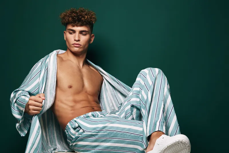 Young man with curly hair wearing striped pajamas and white sneakers sitting against green background. Casual fashion, relaxed pose and confident expression in studio lighting.
