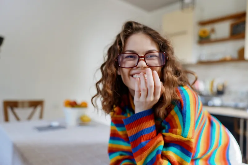 A smiling woman in a colorful striped sweater sits at a bright kitchen table, enjoying a calm moment at home. A candid lifestyle scene capturing warmth, comfort, and everyday happiness.