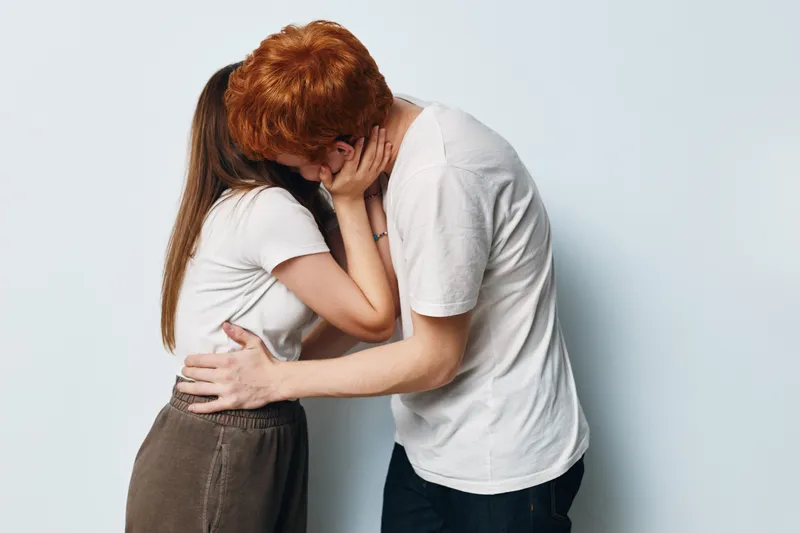 Couple in casual clothing kissing and embracing with hands around neck and waist, showing intimacy and affection against plain white background in studio setting.