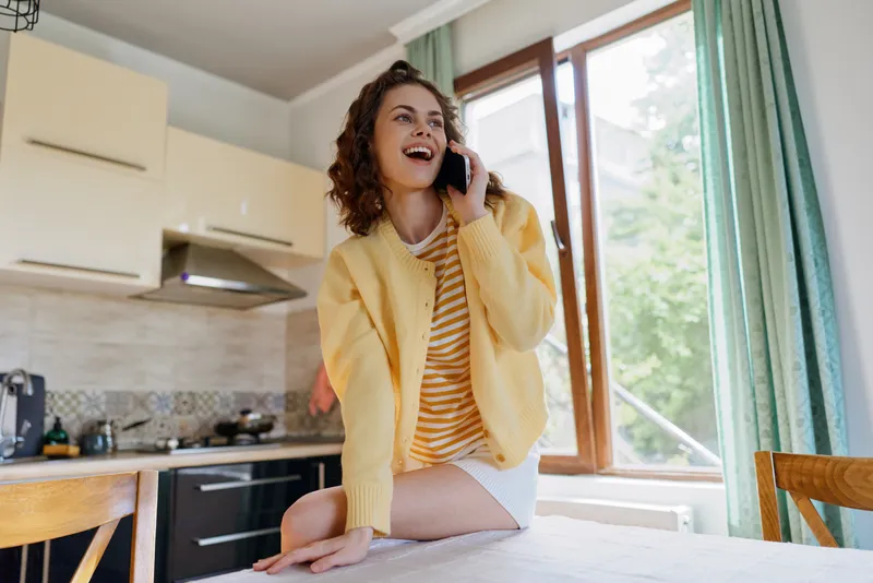 A young woman with curly hair sits on a kitchen counter in a yellow cardigan and striped shirt, talking on a smartphone by a bright window, smiling as daylight fills the open room.