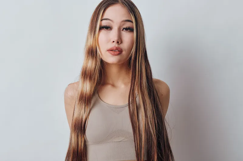 Woman with long straight hair wearing beige tank top looking at camera with neutral expression against plain light background in studio portrait. Female beauty and fashion concept.