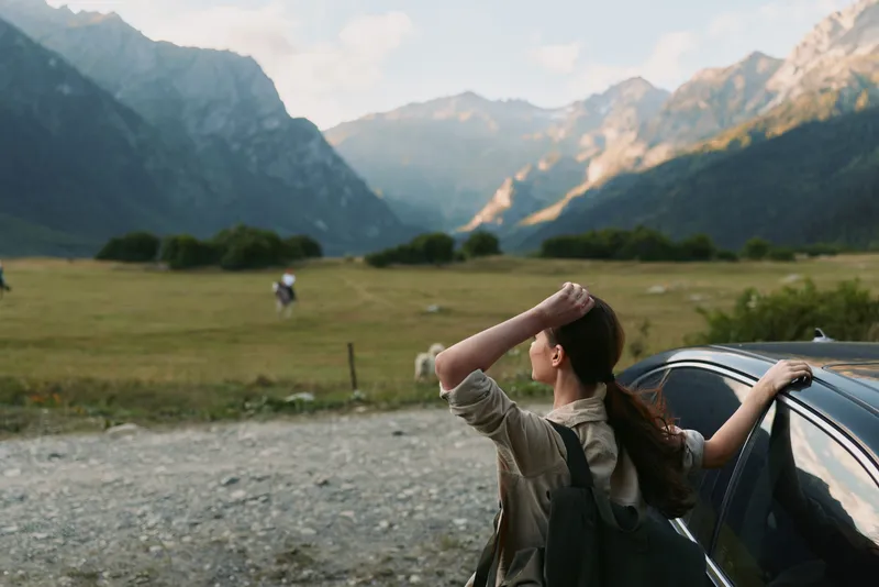 Young woman traveler with backpack stands near car on mountain road looks at scenic landscape enjoys outdoor adventure and peaceful nature view on sunny day.