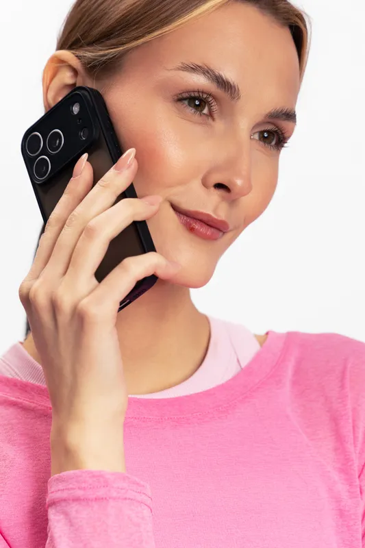 Young woman in pink sweater holds black smartphone near ear and looks away with slight smile. Portrait on white background. Concept for communication, technology, and casual lifestyle.