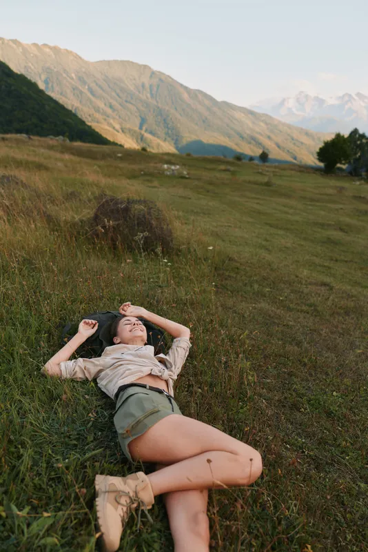 Young woman lying on grass in mountain field, relaxed lifestyle moment, casual outfit, peaceful nature setting, warm sunlight, enjoying outdoor calm