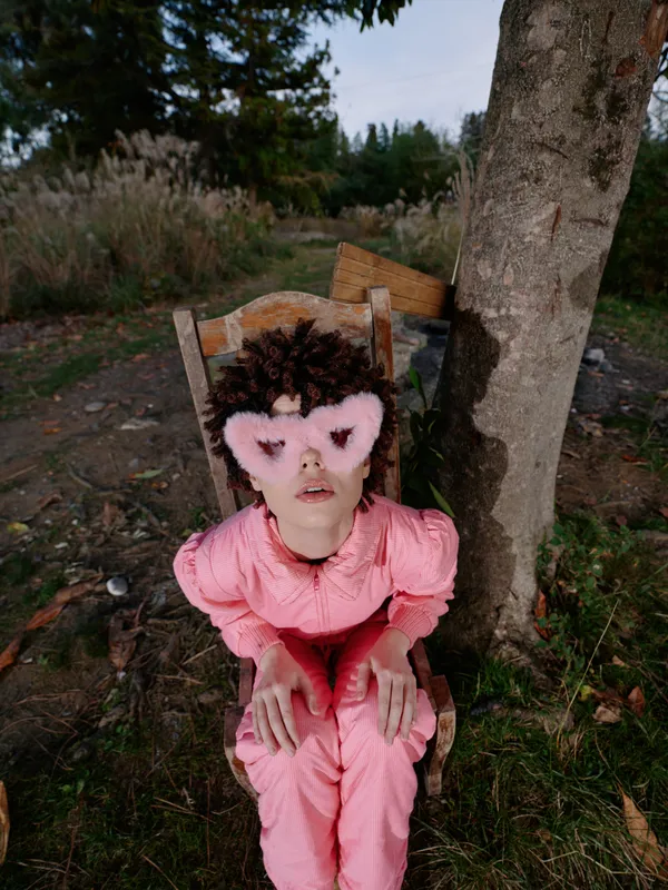 Young child in pink dress sits on a wooden chair outdoors, wearing a heart shaped mask, serene expression, natural setting with trees and plants