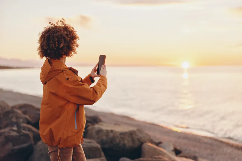 woman on rocky beach at sunset, holding smartphone, orange jacket and curly hair, she watches the ocean horizon, outdoor lifestyle travel moment, warm golden light, gentle breeze