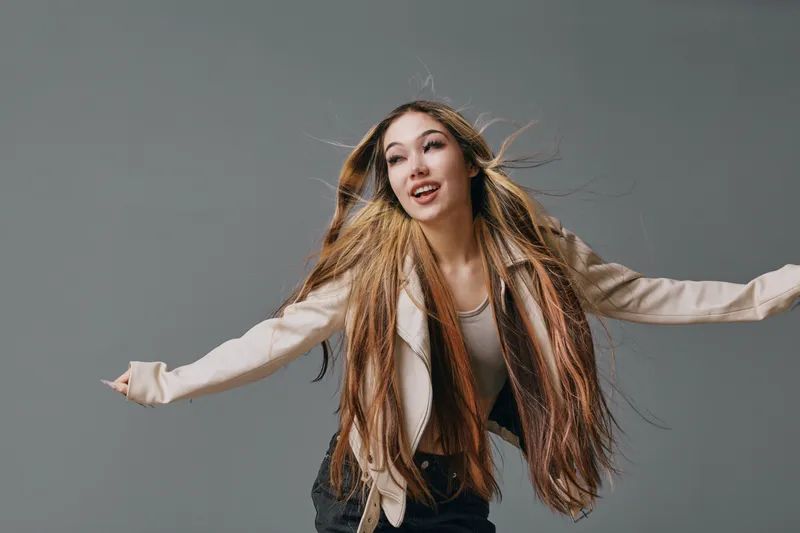 Young woman with long hair wearing light jacket and black pants poses with outstretched arms on gray background. Stylish casual outfit and natural makeup with dynamic hair movement.