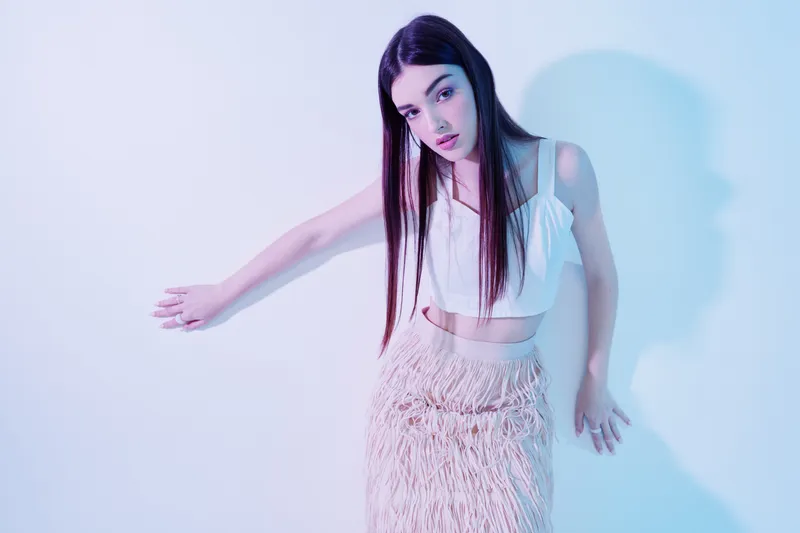 Young woman beauty fashion model poses in a white top and textured skirt against a light background with soft colorful lighting. Concept for style and elegance.