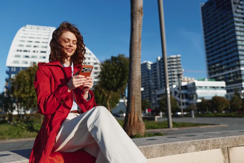 Young woman in red jacket sitting outdoors using smartphone, smiling and enjoying sunny day in urban park with modern buildings in background.