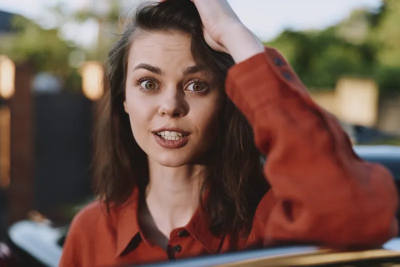 Young woman with brown hair in a rust shirt adjusts her hair outdoors, a candid moment that conveys casual confidence, everyday style, and a relaxed mood