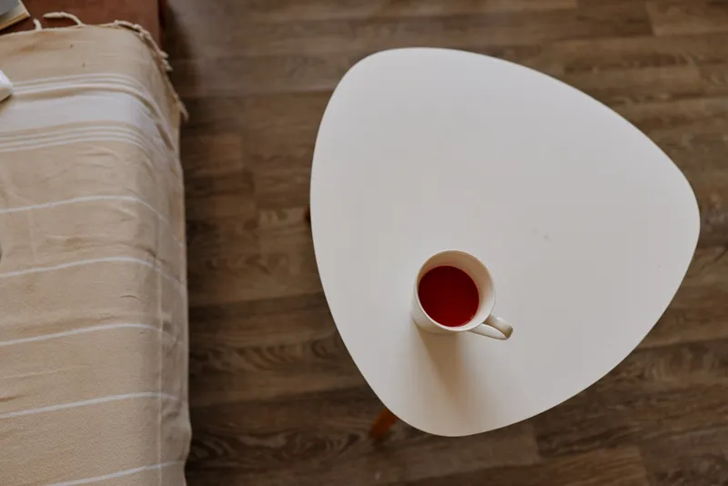 Cozy home scene with a white triangular table and a red coffee cup, wooden floor, natural lighting, simple interior, inviting calm atmosphere, a quiet moment of relaxation and everyday comfort