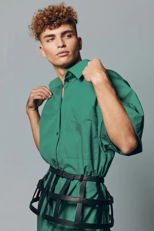 Young man with curly hair wearing green shirt and black harness posing against gray background in studio. Fashion model with confident expression and stylish outfit for editorial look.
