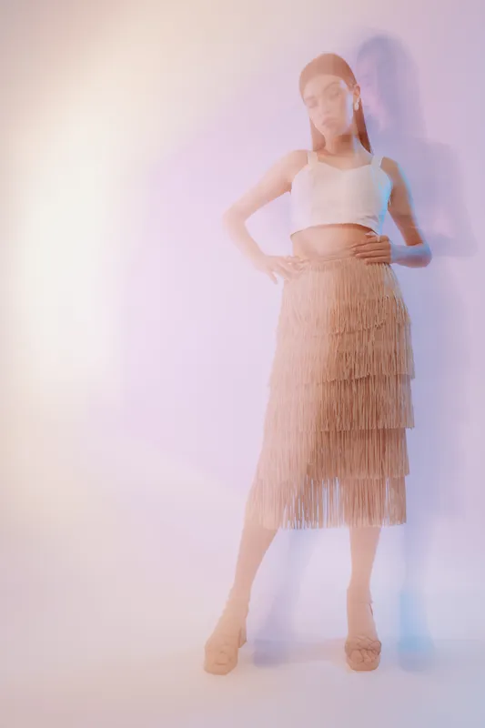 Fashionable woman in a beige fringe skirt and white crop top poses confidently against a light background. Soft lighting highlights beauty, style, and modern fashion concept.