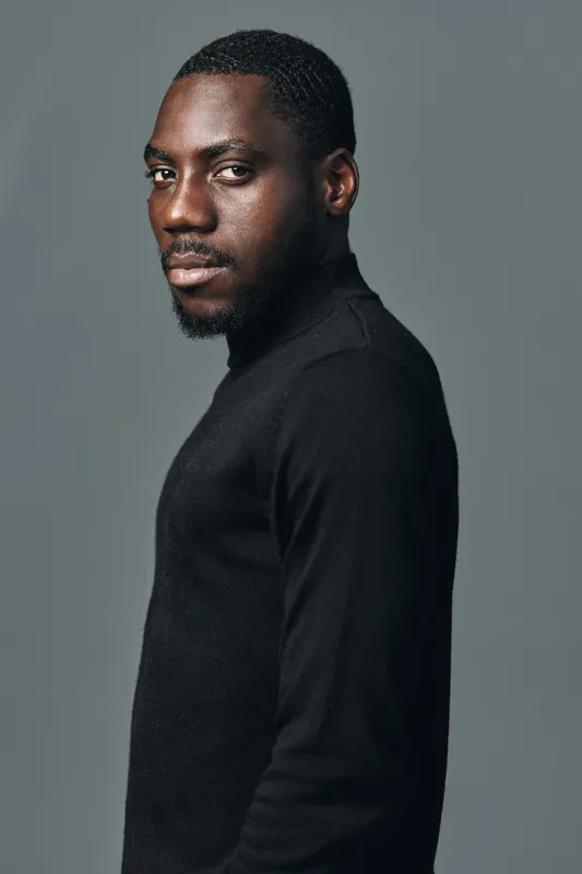 African man portrait in creative conceptual visual style wearing black turtleneck sweater against gray background. Confident male model with dark skin and short hair looking at camera in studio.