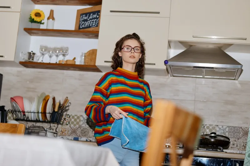 Young woman with curly hair and glasses stands in a bright kitchen wearing a colorful striped sweater, folding a blue denim garment with a focused calm attitude.