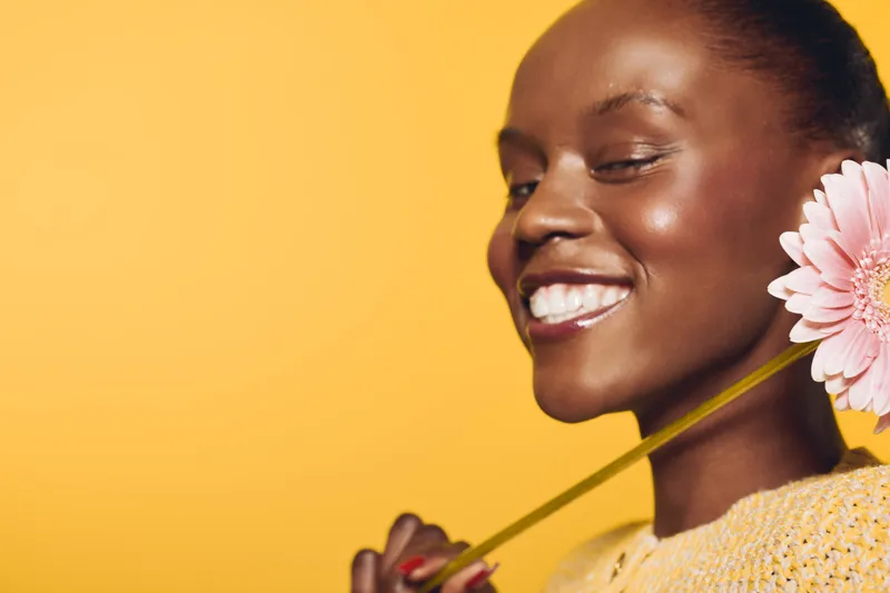 Smiling African woman with dark skin holds pink flower near face, wearing yellow sweater on yellow background. Concept of happiness, natural beauty, wellness, and floral skincare.