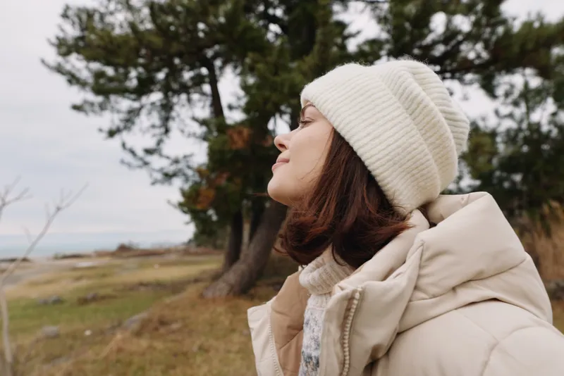Woman portrait in outdoor setting with a coat and beanie, standing near a coastal landscape that conveys calm, cold weather and a quiet autumn mood.