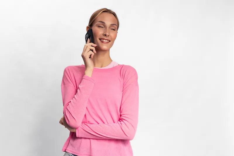 Young woman in pink sweater talking on phone with eyes closed smiling, isolated on white background. Concept of communication, happiness, casual lifestyle and technology use.