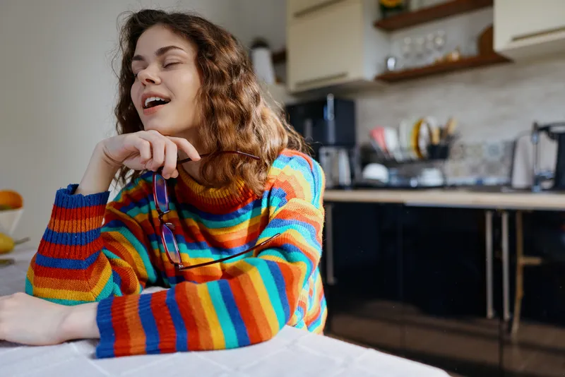 Young woman sits at a kitchen table at home, wearing a colorful rainbow striped sweater, relaxed pose, smiling, casual lifestyle shot in a cozy domestic setting with soft light