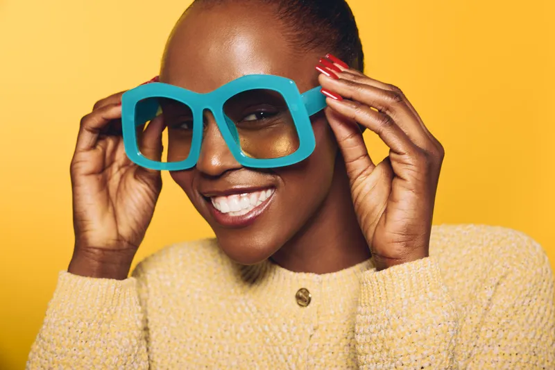 Smiling African American woman with dark skin wears oversized blue sunglasses and yellow sweater. Close-up on bright yellow background. Concept of fashion, beauty, and joyful mood.