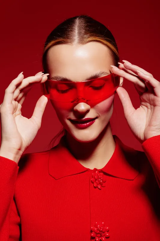 Young woman in red sweater adjusts red translucent glasses on red background. Fashion portrait with stylish accessories and confident expression. Concept of modern style and bold colors.