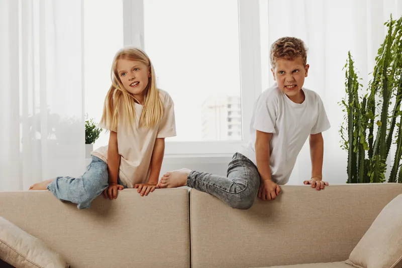 Two children sitting playfully on a beige sofa in a bright living room with plants near the window, enjoying a relaxed moment.