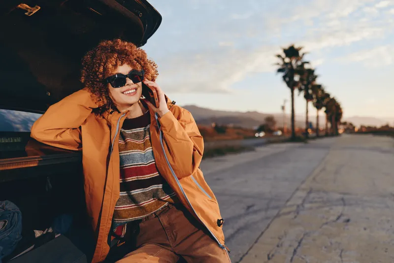 Cheerful woman in sunglasses by a car, smiling and posing outdoors, curly hair and warm fashion coat, sunset road background, relaxed lifestyle travel mood, golden natural light