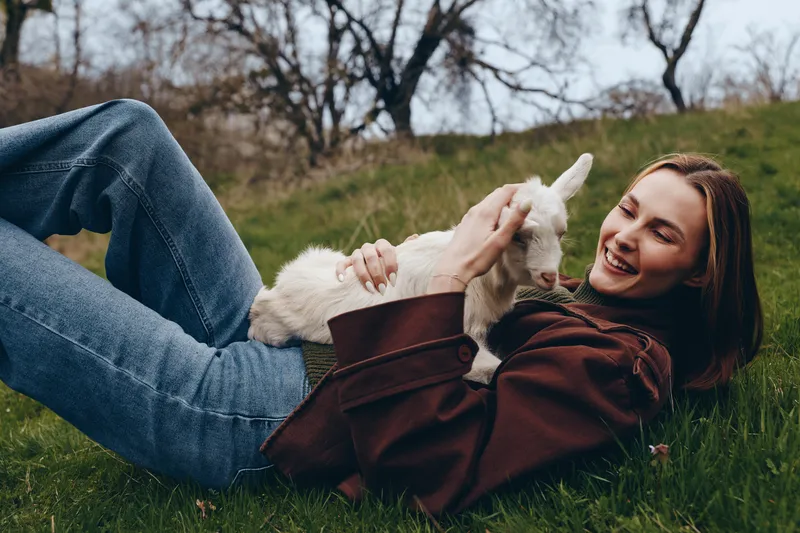 A woman lying on grass outdoors, cuddling a small white goat kid, smiling with contentment. A lifestyle moment showing bonding with animals, relaxation, and rural joy.