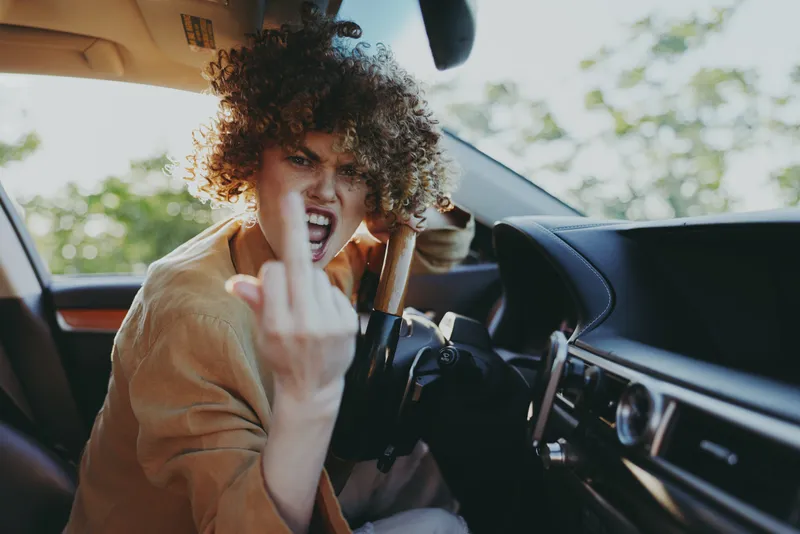 Young woman with curly hair sits inside a car, flashing a rebellious gesture. Casual style, sunny interior, retro film colors convey bold attitude and carefree road trip energy.