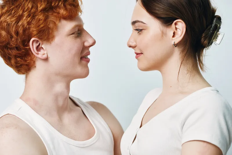 Young man and woman looking at each other with smiles in casual light clothing on white background. Portrait of happy couple in closeup with natural expressions and soft lighting.
